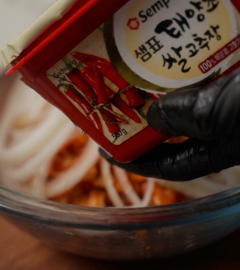 A hand in a black glove holding a container of red chili paste over a mixing bowl to add a scoop of paste.