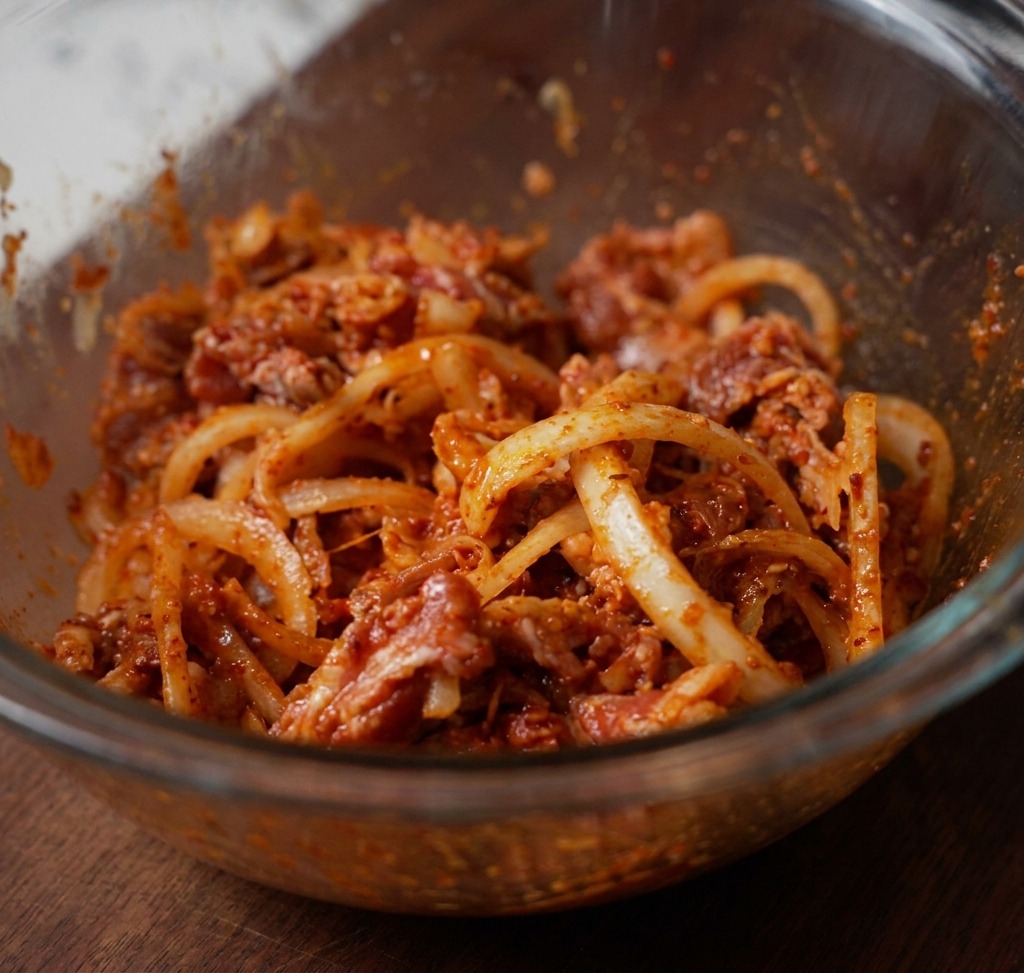 Thinly sliced beef, white onions, and red chili paste thoroughly combined in a clear glass bowl, ready for marinating.