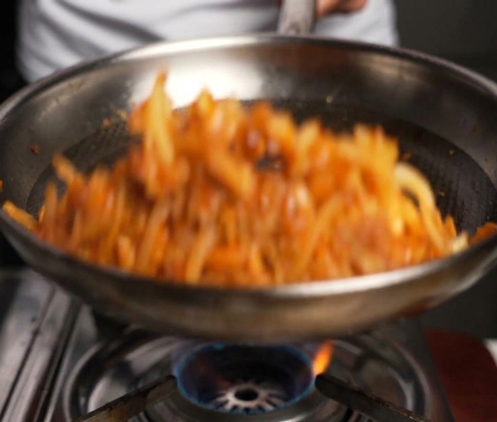 Beef and onions being tossed and stir-fried in a metal pan with a wooden spatula over a gas flame.
