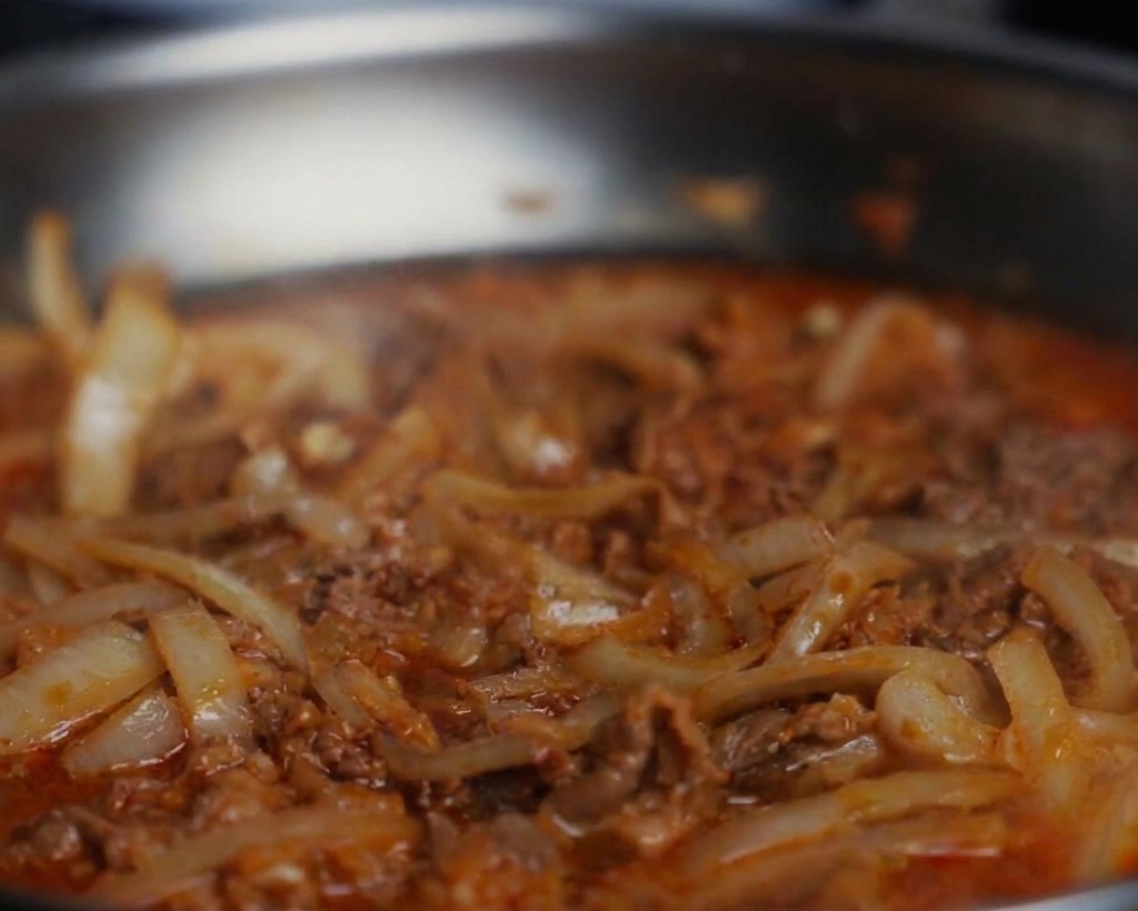 Close-up of black pepper and soy sauce being added to a simmering pan of beef and onions.