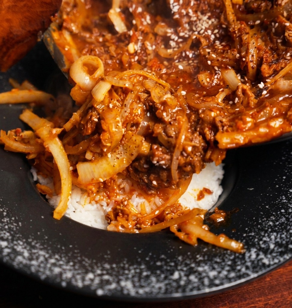 Rich beef and onion stew being poured from a pan onto a bed of steamed white rice in a black bowl.
