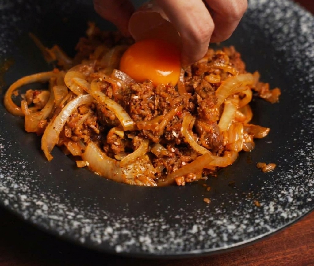 A hand using an eggshell to gently drop a vibrant orange egg yolk onto a mound of savory stir-fried beef and onions.