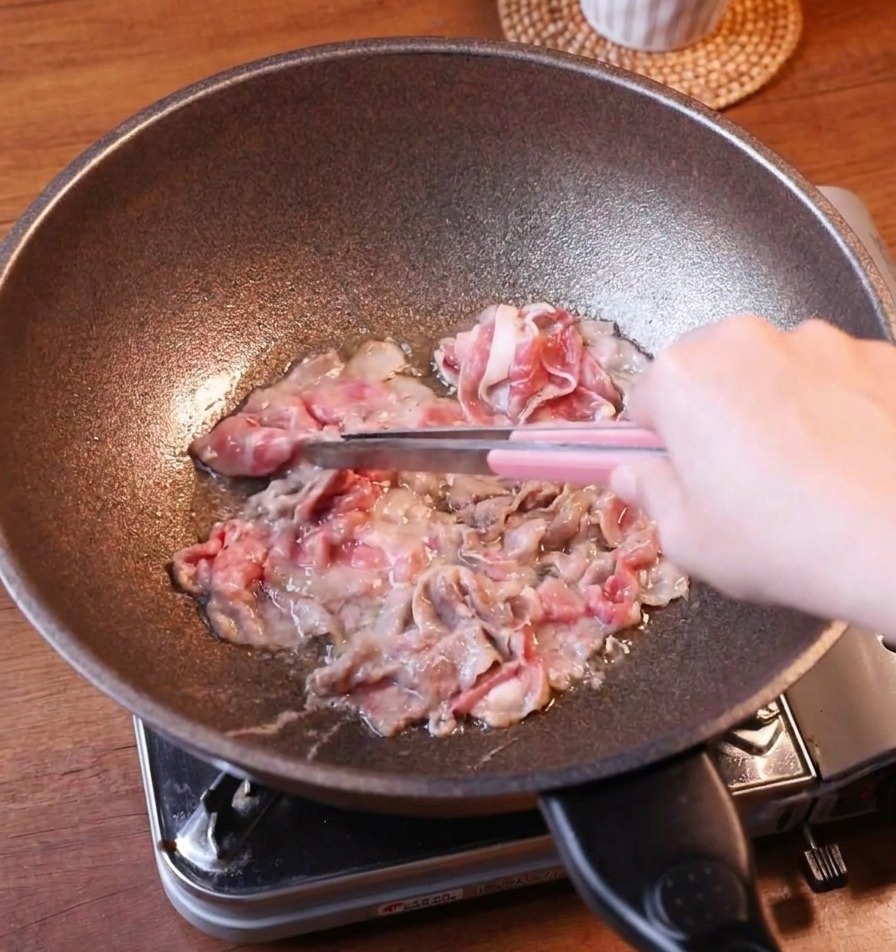 Thin slices of browned fatty beef being lifted from a dark skillet with pink-handled metal tongs, showing the meats curled texture.