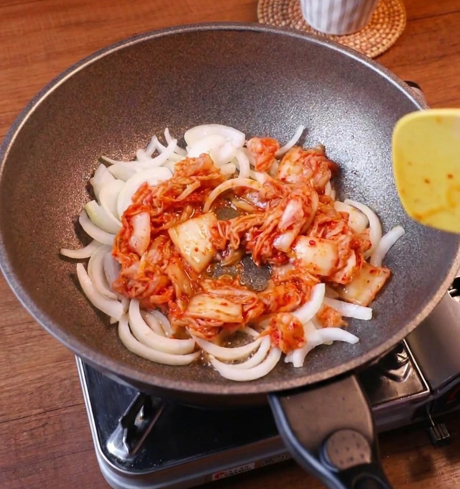 Sliced white onions and vibrant red kimchi being stir-fried together in a pan with a yellow spatula.