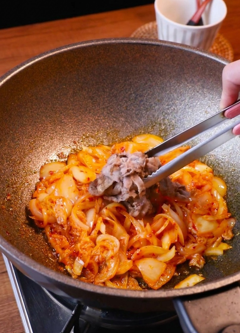 Tongs returning browned beef slices to a pan filled with sautéed kimchi and onions.