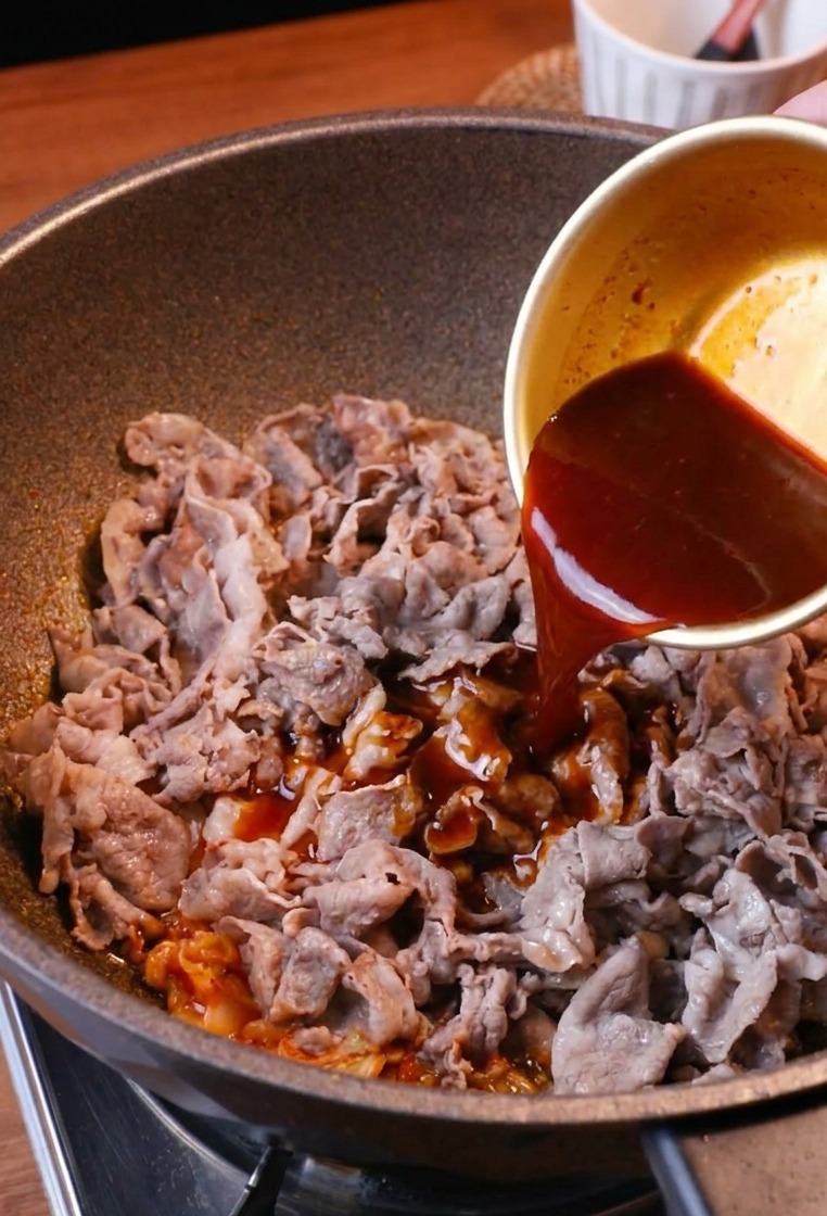 Savory reddish-brown sauce being drizzled from a bowl over a pan of stir-fried beef and kimchi.