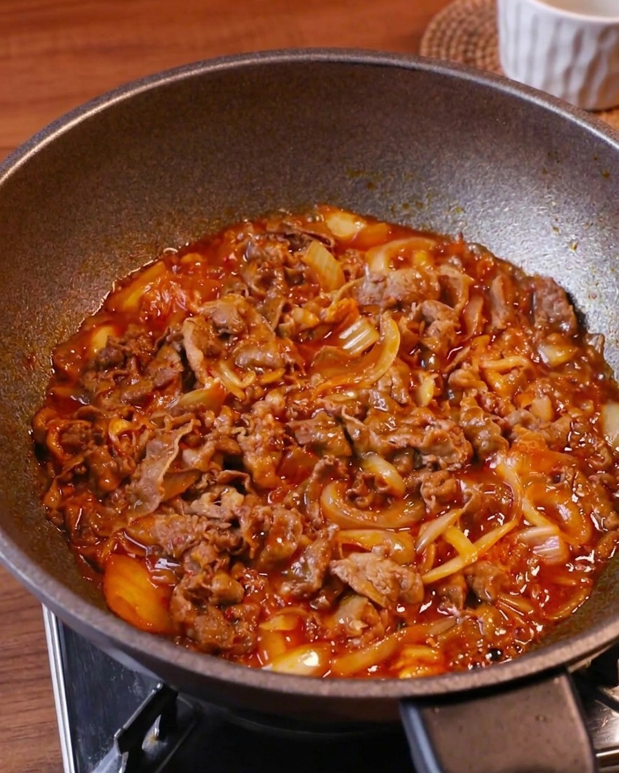 Tender beef slices, kimchi, and onions simmering in a rich, bubbling red sauce in a wok under high heat.