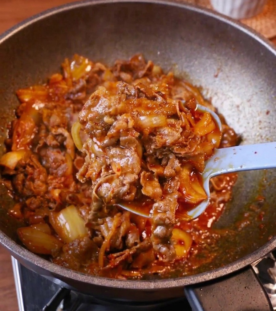 A light blue spoon layering a generous portion of glazed kimchi beef onto a bowl of steaming white rice.