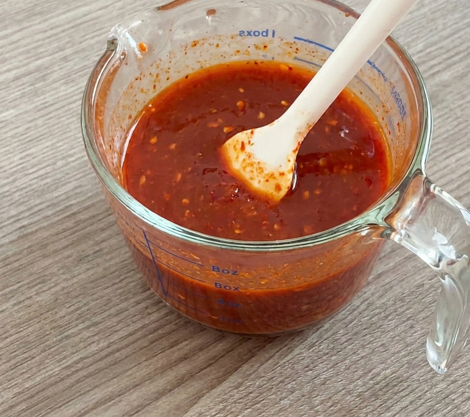 A white silicone spatula stirring a thick, bright red sauce mixture inside a glass measuring cup.
