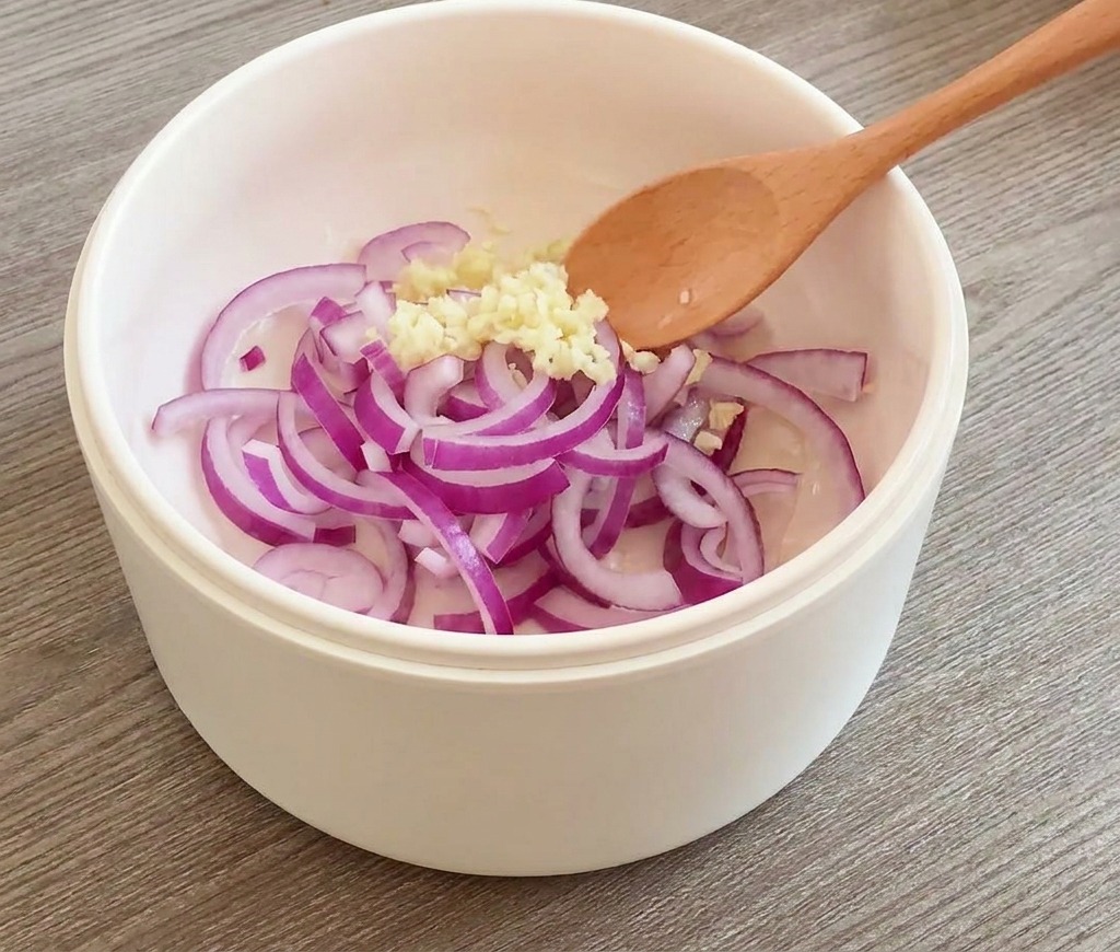 Red onion rings and minced garlic being stirred with a wooden spoon in a white pan.