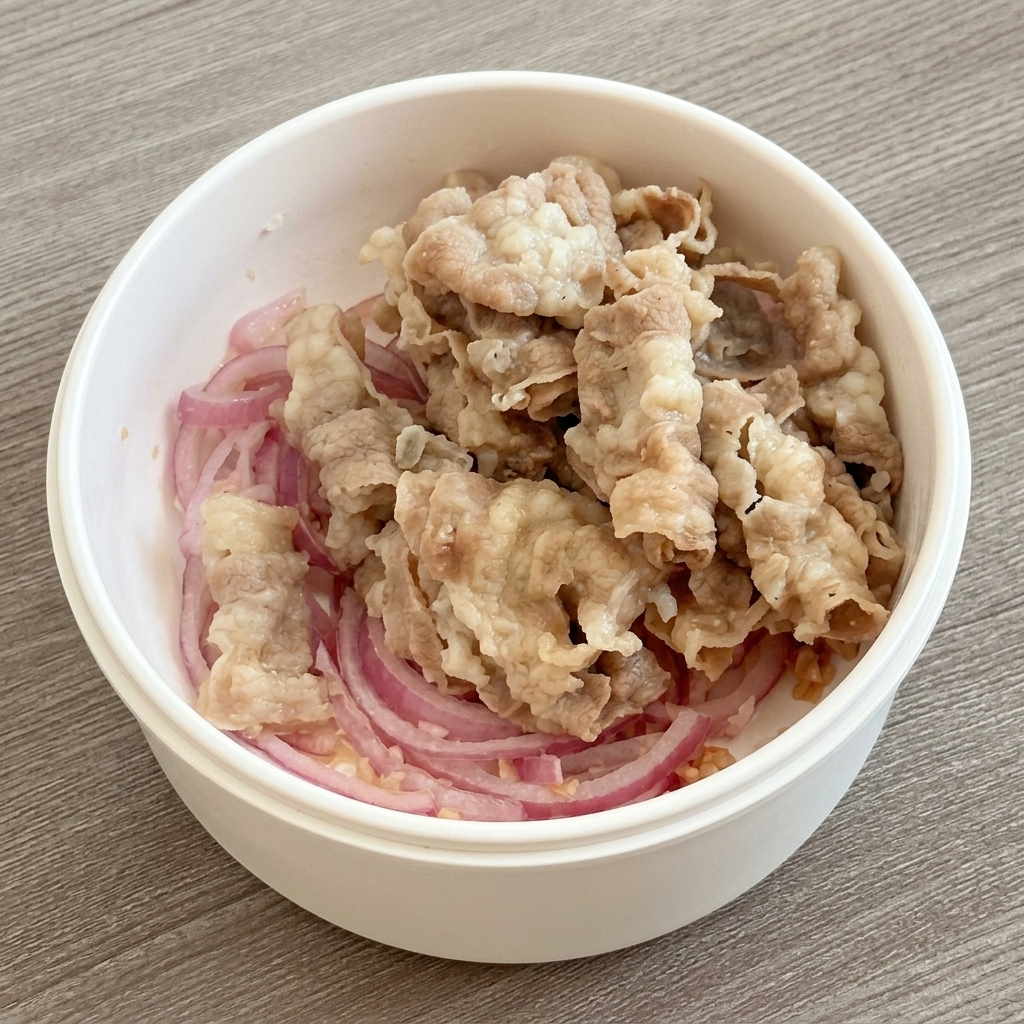 Blanched beef slices placed on top of sliced red onions in a white cooking vessel.