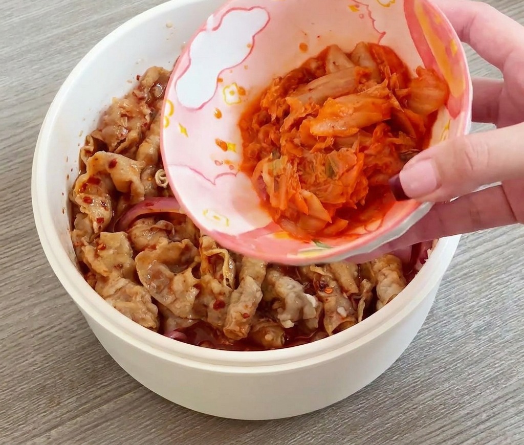 A hand pouring a small bowl of sliced red kimchi into a pan of stir-frying beef and onions.