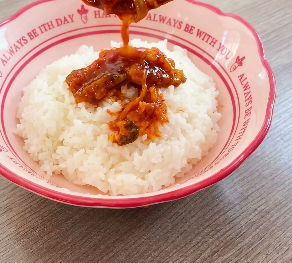 Savory red kimchi and beef mixture being poured from a spoon onto a mound of white rice in a decorative pink bowl.