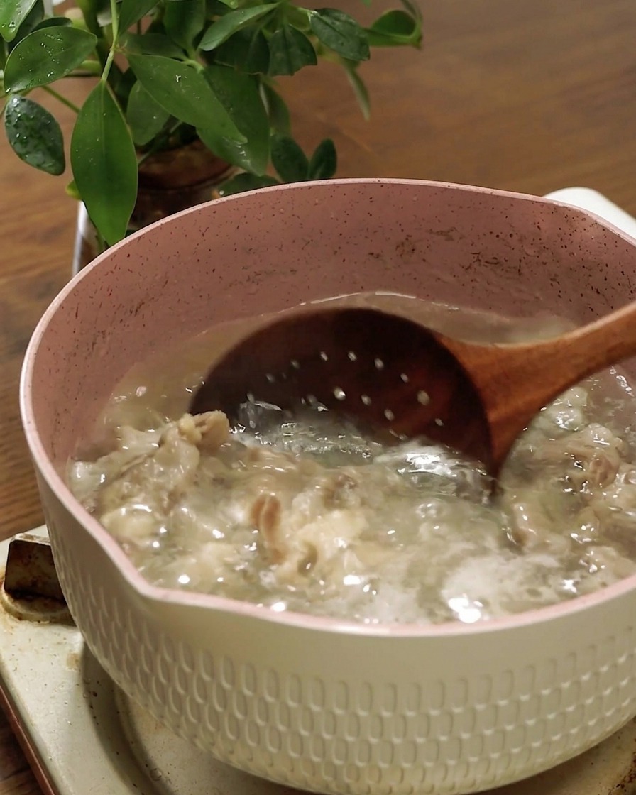 A wooden slotted spoon lifting cooked fatty beef out of a pot of boiling water.