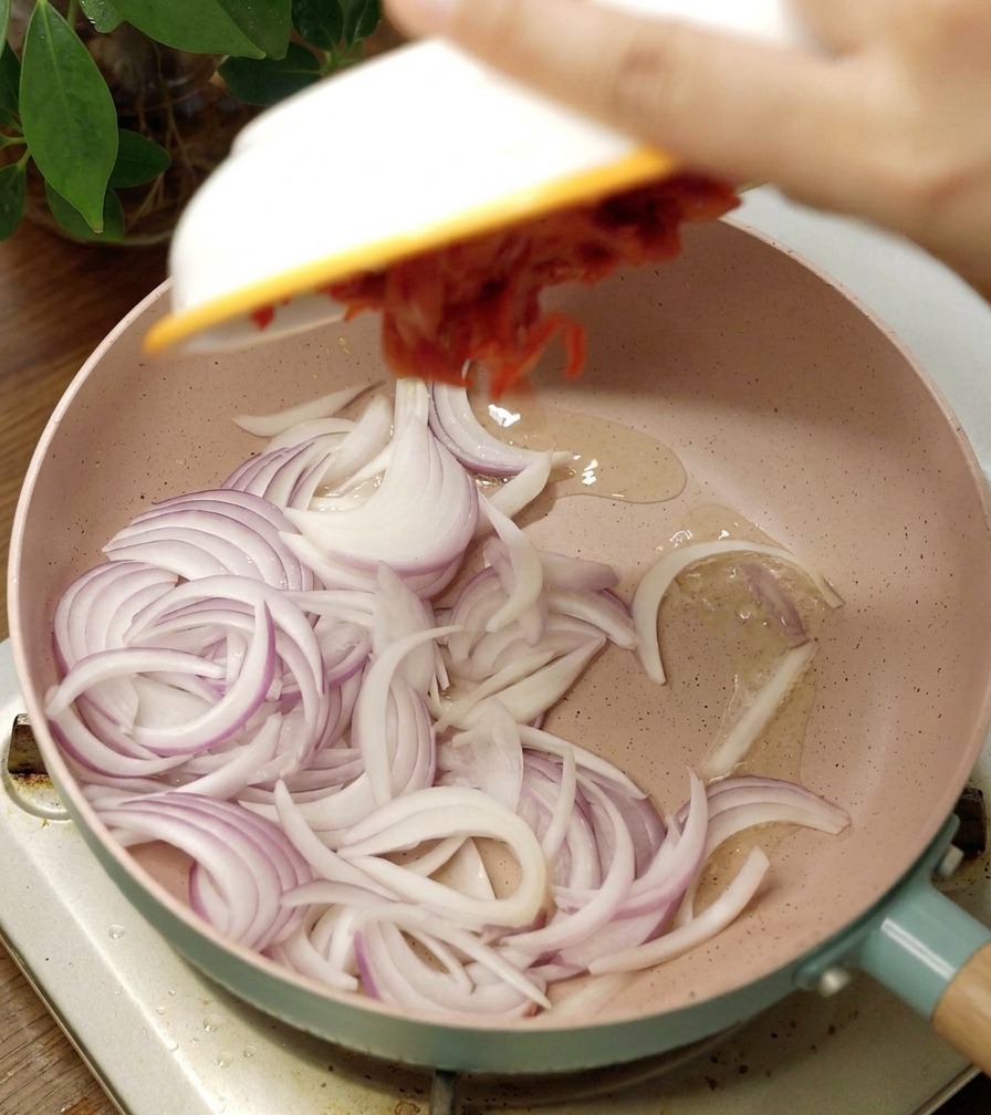 A pink ceramic pan containing sliced raw red onions and a scoop of spicy red kimchi being added from a white bowl.