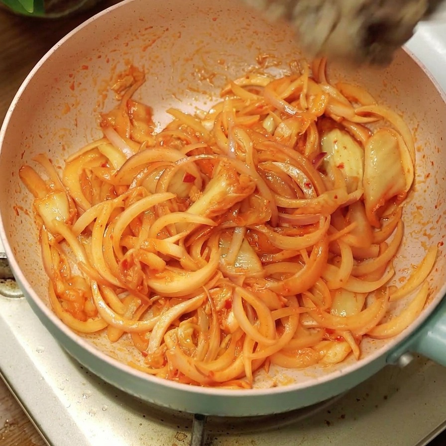 Cooked beef slices being poured from a white bowl into a pink frying pan containing stir-fried onions and kimchi.