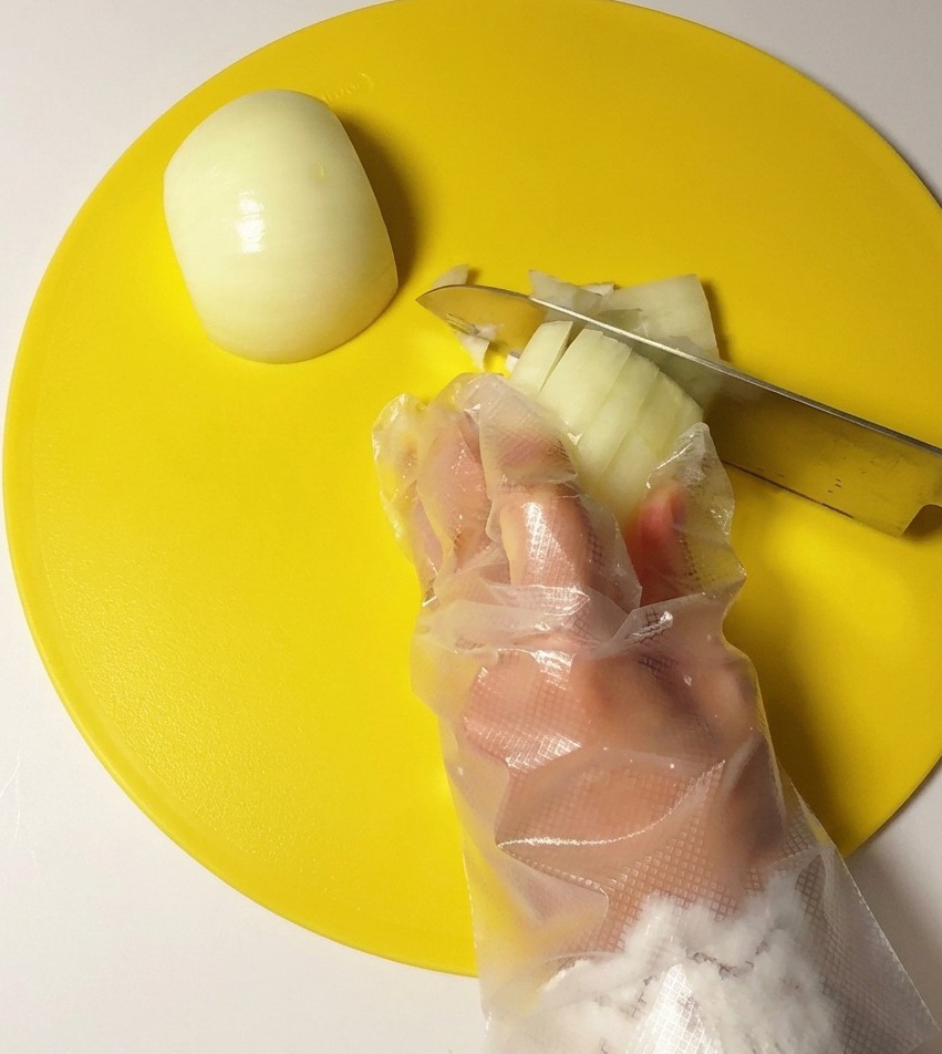 A hand wearing a clear plastic glove dicing a white onion with a stainless steel knife on a bright yellow circular cutting board.