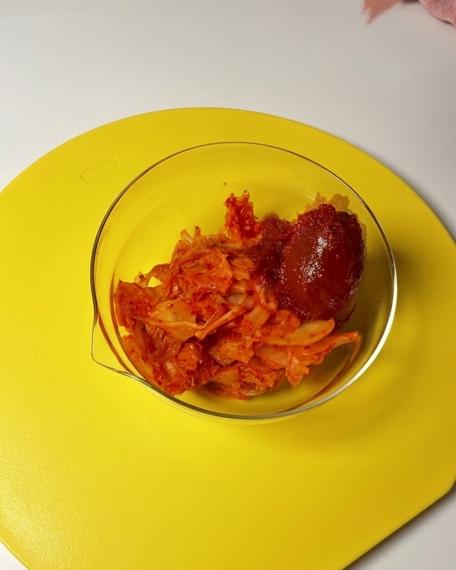 A spoonful of red gochujang and chili powder being added to a glass bowl filled with chopped kimchi.