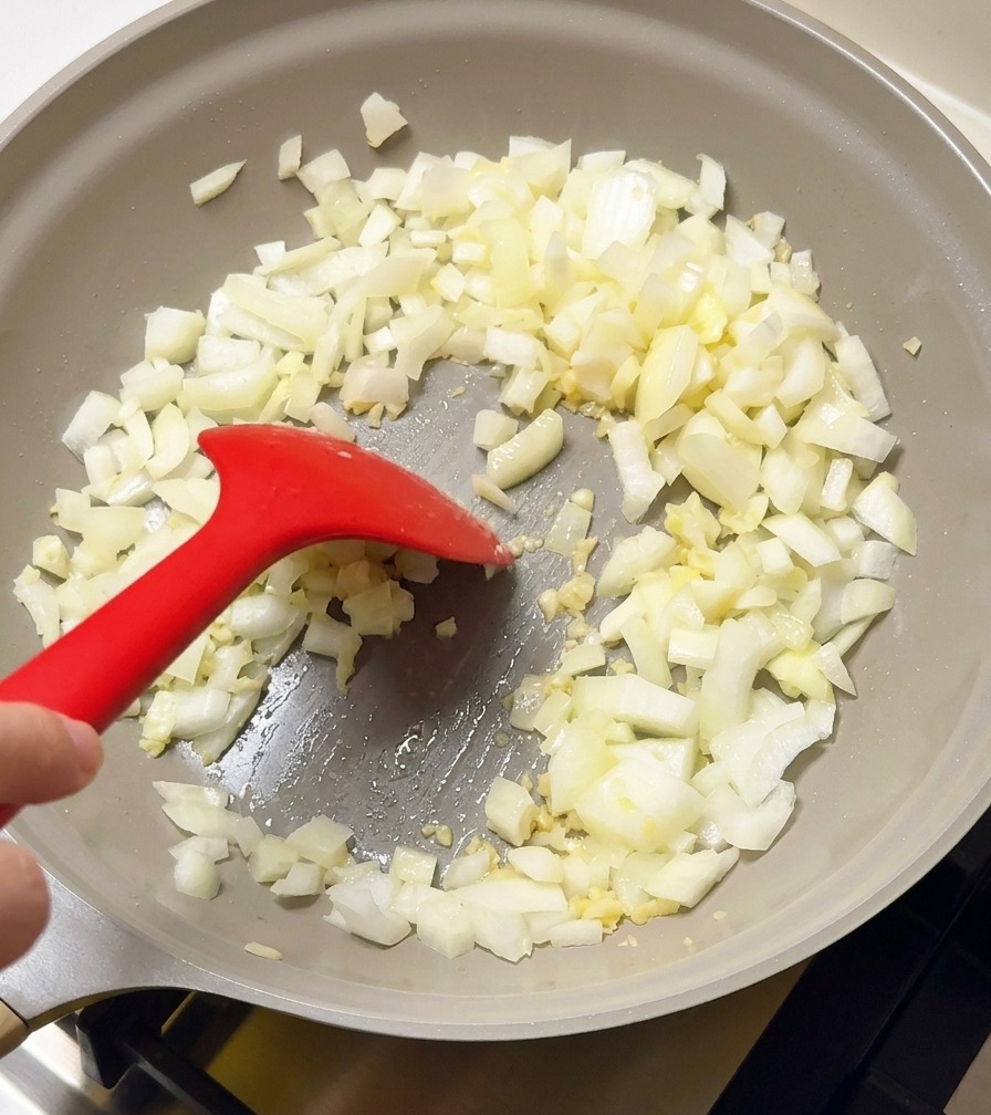 Diced white onions and garlic being sautéed in a light gray frying pan with a red silicone spatula.