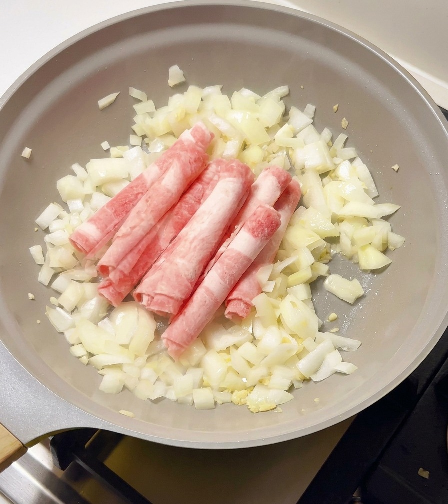 Thinly sliced, frozen raw beef rolls being added to a pan with sautéed white onions.