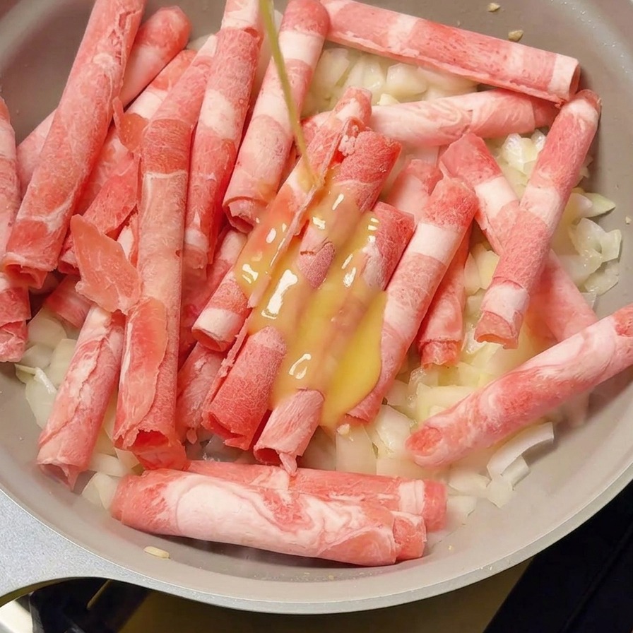 Yellow ginger juice being poured from a small squeeze bottle onto frozen beef rolls layered over white onions in a grey pan.