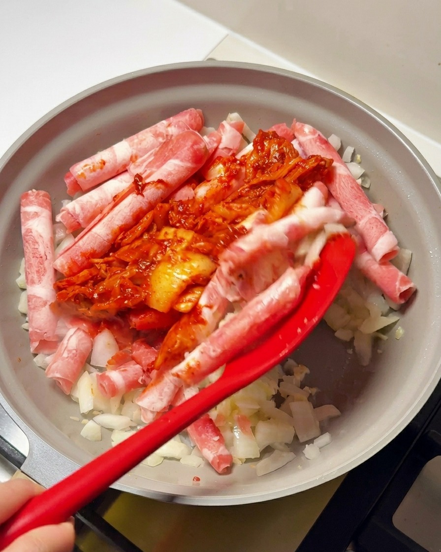 A red spatula adding a chunky red kimchi and chili paste sauce onto beef rolls and chopped onions in a frying pan.