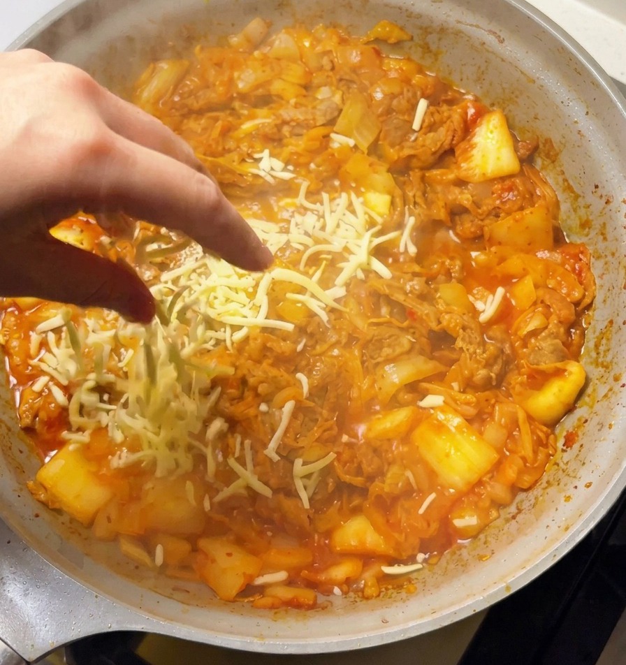 A hand sprinkling a generous amount of shredded white mozzarella cheese over a bubbling pan of spicy kimchi and beef stir-fry.