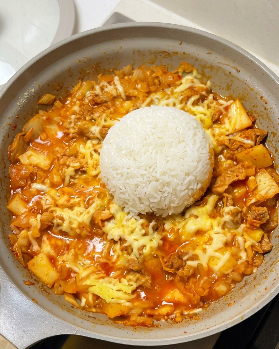 A perfectly round dome of white cooked rice placed in the center of a frying pan, surrounded by melted cheese and a red kimchi beef base.