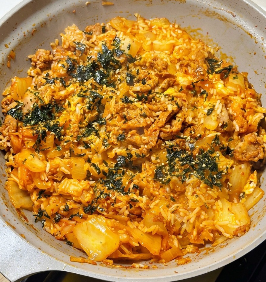 A hand sprinkling dark green roasted seaweed flakes over a steaming pan of finished kimchi beef fried rice with visible cheese pulls.