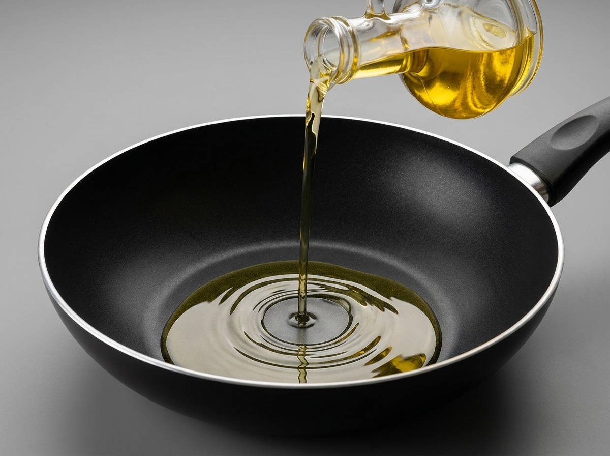 Clear cooking oil being poured from a glass bottle into a clean, black non-stick wok.