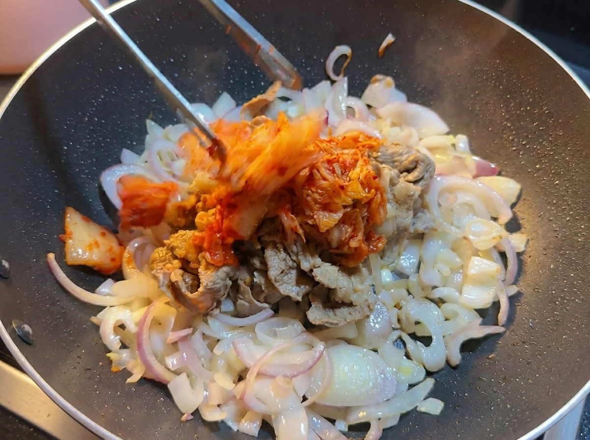 Chopped cabbage kimchi being added with tongs into a wok containing sliced beef and onions.