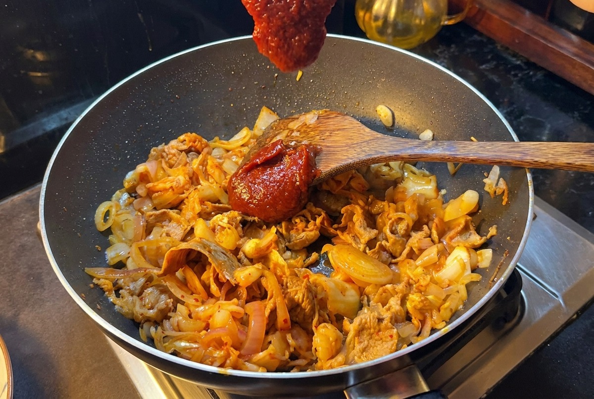 A wooden spatula holding a large scoop of thick red Korean chili paste over a pan filled with stir-fried beef slices, onions, and kimchi.