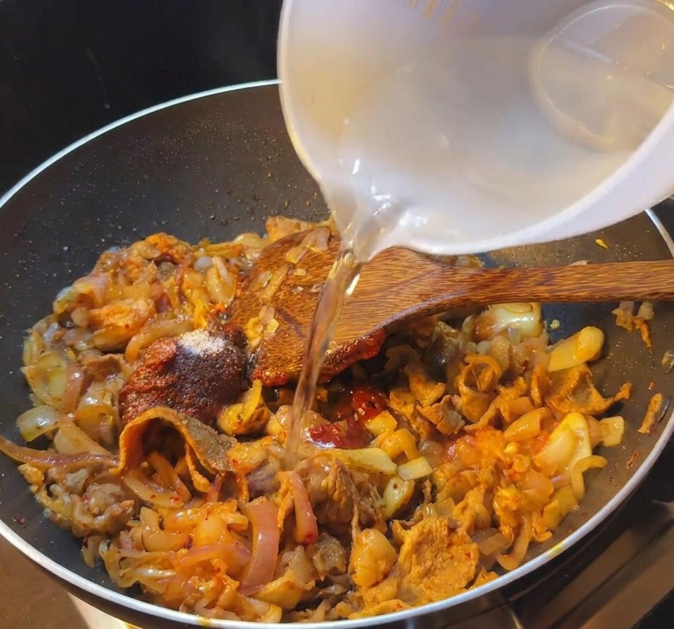 A person pouring clear water from a white bowl into a black frying pan containing sliced beef and red spicy kimchi sauce.
