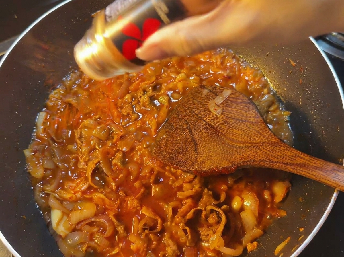 Red chili powder being shaken from a black container into a pan of bubbling red sauce with beef and onions.