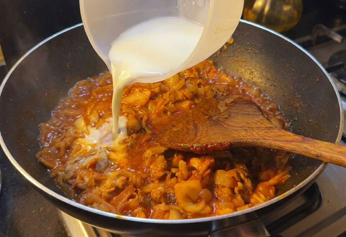 Milky white starch water being poured from a plastic cup into a pan of simmering beef and kimchi in a rich red sauce.