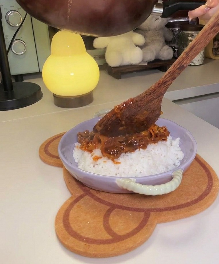 A pan being tilted to pour a thick, red beef and kimchi mixture onto a bed of fluffy white rice in a lavender-colored bowl.
