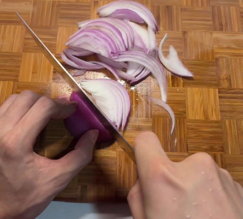 A person slicing a fresh red onion into thin, even strips on a wooden cutting board with a sharp chefs knife.