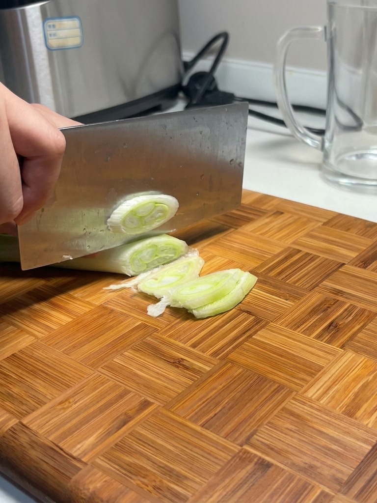 A large cleaver slicing through a thick leek on a wooden cutting board, creating several round white and green slices.