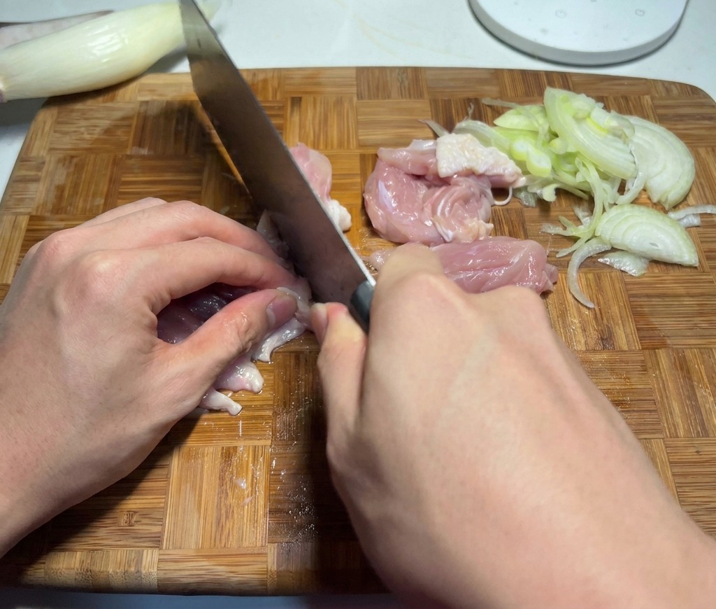 A chef cutting raw pink chicken thigh meat into small bite-sized pieces on a cutting board next to some sliced vegetables.
