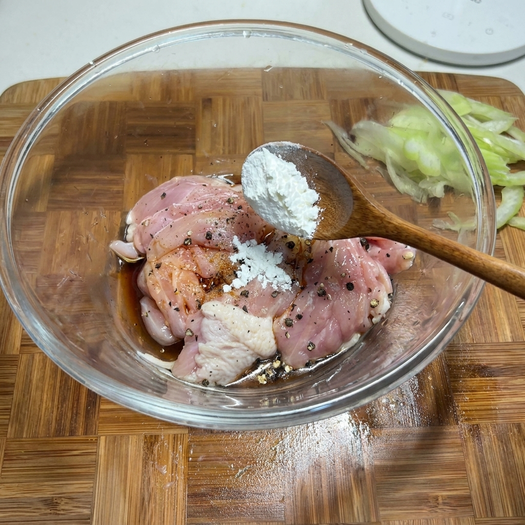 A wooden spoon adding white cornstarch to a glass bowl containing seasoned raw chicken pieces.