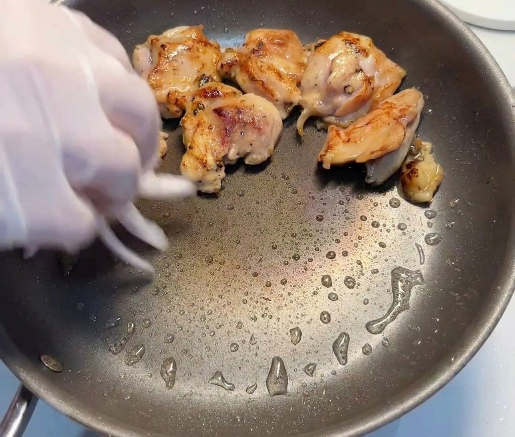 Slices of red onion being added by a gloved hand into a pan containing seared chicken thigh pieces.
