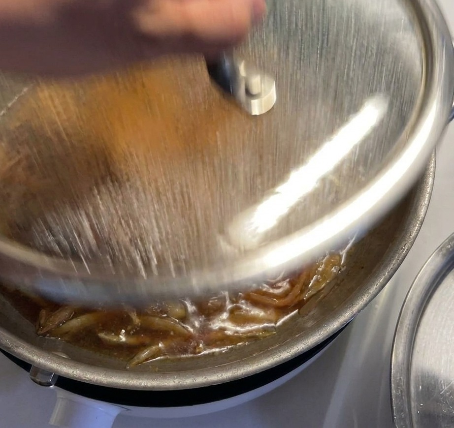 A hand placing a glass lid with a silver handle onto a pan where chicken and onions are simmering in a bubbling dark sauce.
