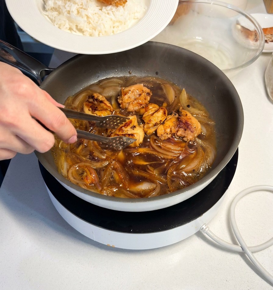 Metal tongs placing a piece of succulent, sauce-coated chicken onto a mound of fluffy white rice in a white ceramic dish.