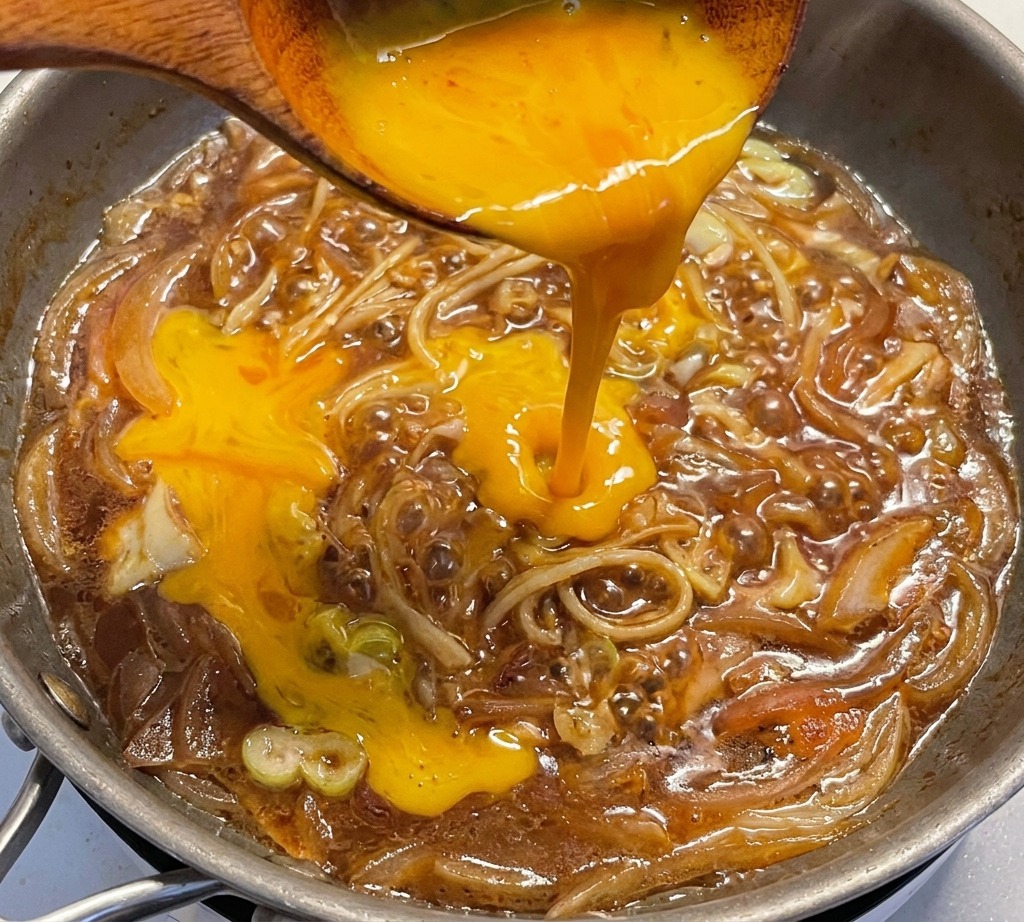 Beaten egg yolks being poured from a wooden ladle into a skillet filled with a savory onion and leek mixture simmering in sauce.