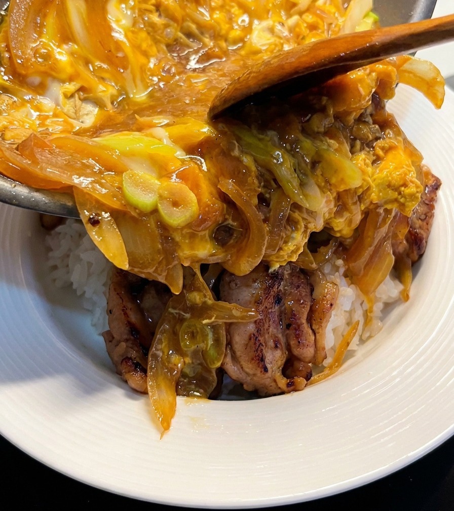 A wooden spoon helping slide a glossy mixture of cooked eggs and onions from a pan onto a bowl of white rice topped with browned chicken pieces.