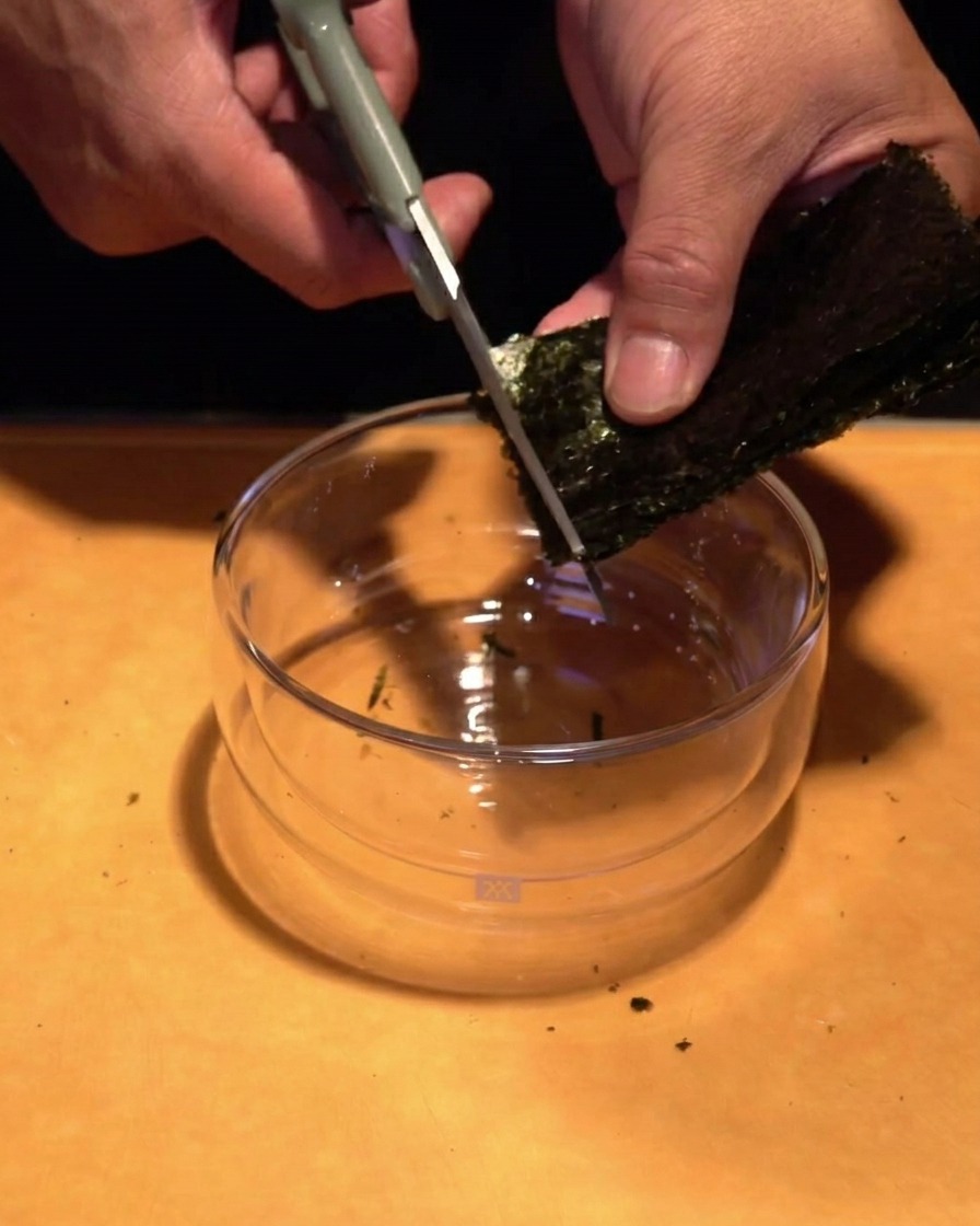 Hands using kitchen scissors to cut a stack of dark green seaweed into thin matchstick pieces over a clear glass bowl.