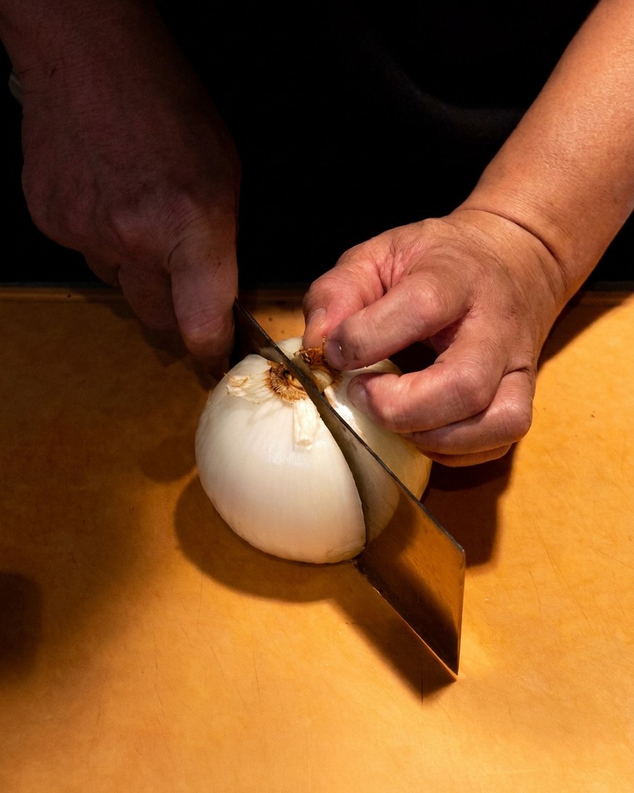 A close-up of hands carefully slicing a white onion into thin wedges on a wooden board.