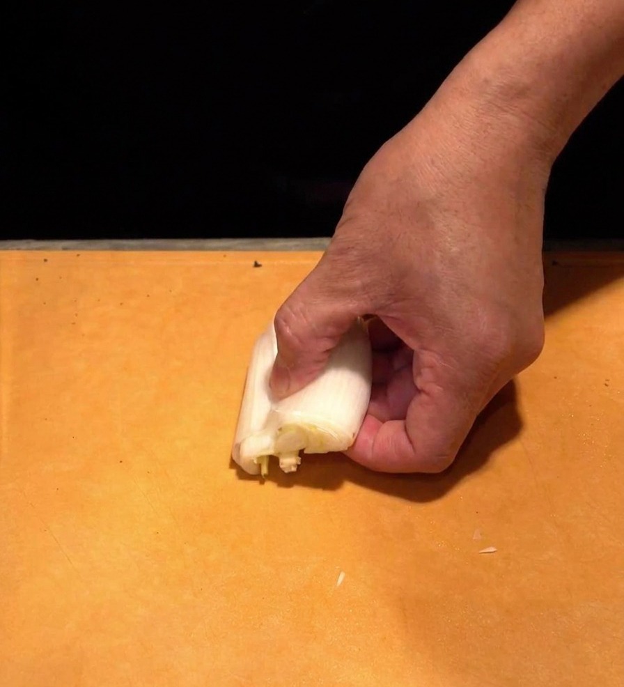 A hand holding a thick white section of a Japanese naganegi onion on a cutting board, slicing it into paper-thin white strips.