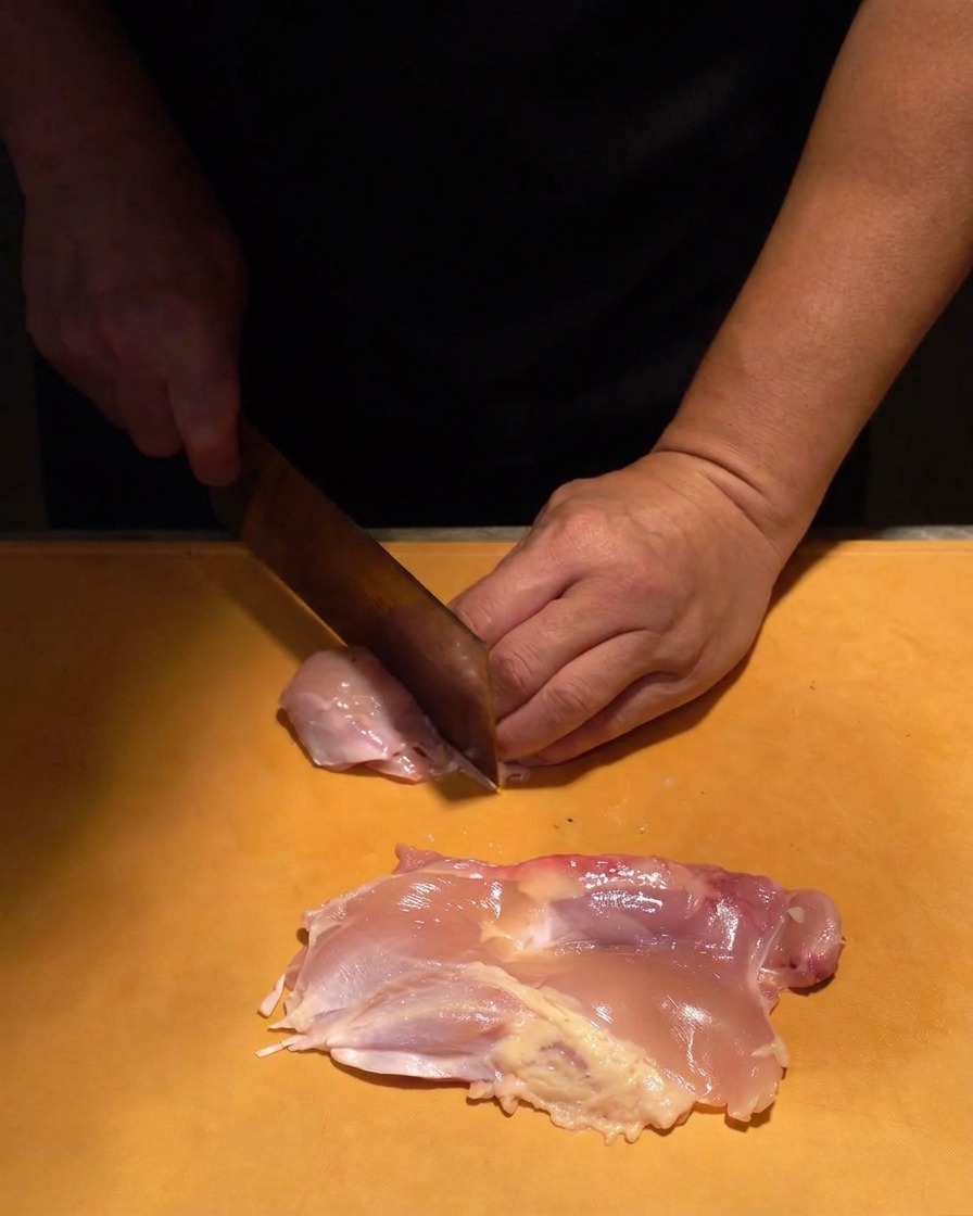 Raw pink chicken thigh meat being diced into uniform pieces with a sharp knife on a textured orange board.