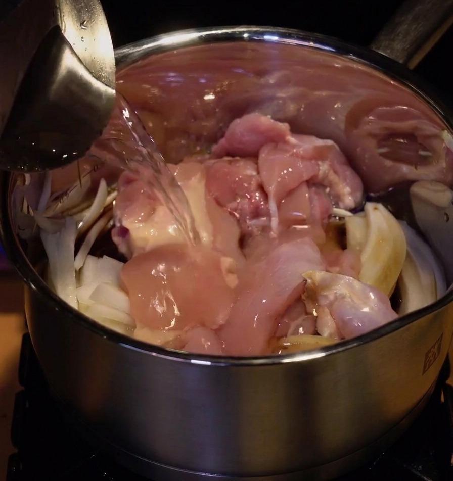 Water being poured from a metal pitcher into a stainless steel pot filled with raw pink chicken thighs and sliced white onions.